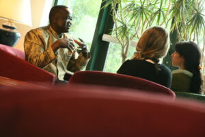 In a lounge setting, sitting in a circle on upolstered chairs, a professor and two students in a discussion